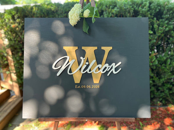 Last name sign with monogram and established date, on a wooden background with customizable colors - example on a black background, gold monogram, white text, gold date