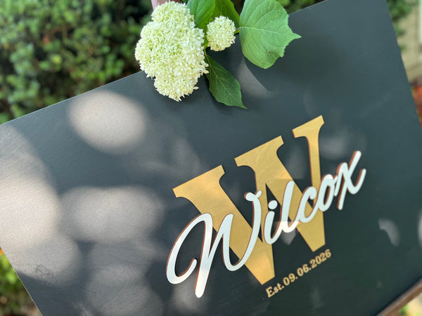 Last name sign with monogram and established date, on a wooden background with customizable colors - example on a black background, gold monogram, white text, gold date