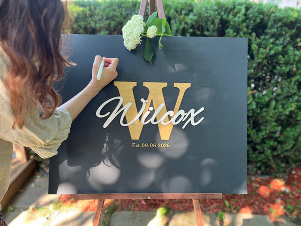 Last name sign with monogram and established date, on a wooden background with customizable colors - example on a black background, gold monogram, white text, gold date