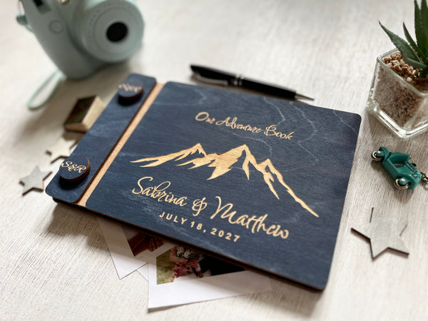 A personalized dnavy blue wedding guest book with mountain engraving and personalized names, styled on a light wood surface with a pen, succulent, and small decorative items.