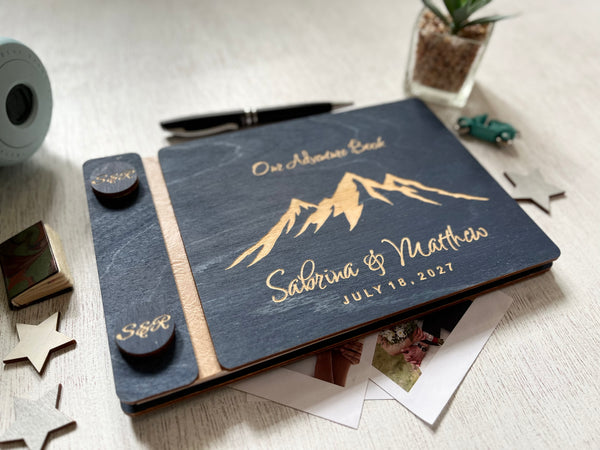 A personalized navvy blue wedding guest book with laser-engraved mountain graphics and names, styled on a light wood tabletop with a pen, plant, and star decorations.