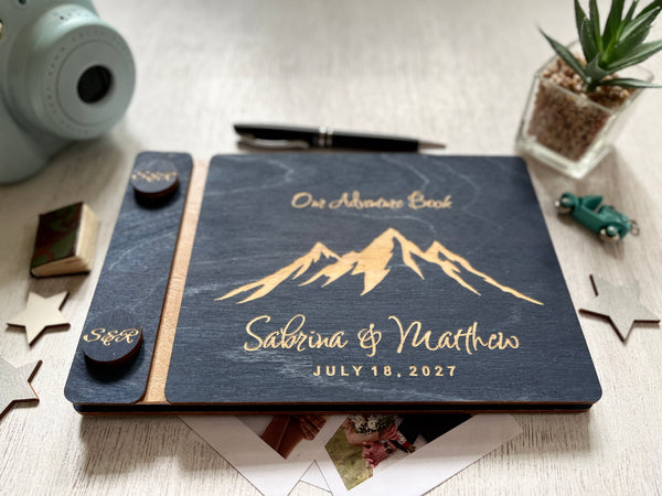 A personalized mountain-themed keepsake book with a navy blue wooden cover and laser-engraved text reading 'Our Adventure Book' and personalized names with a personalized date also included. The book is styled on a light wood tabletop with a pen, small plant, star-shaped cards, and other decorative props around it.