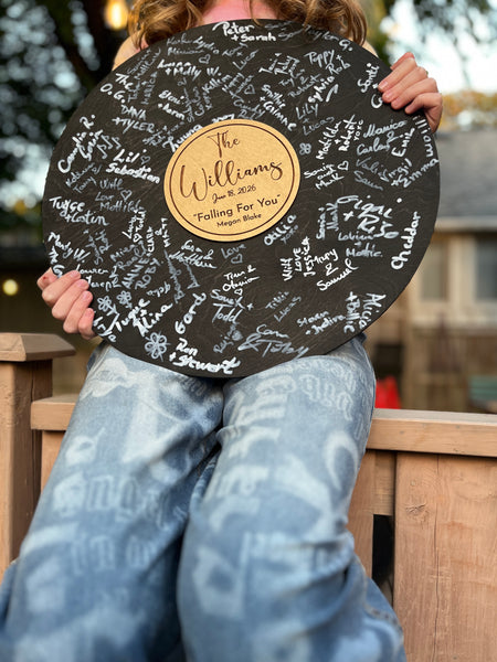 Person holding a circular vinyl record black board with gold center and engraved text, sitting outdoors.