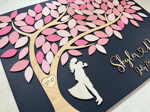 side detail of wedding guest book alternative with tree of life and pink shades for the leaves and embracing couple underneath the tree that has a carved heart with the couple's initials