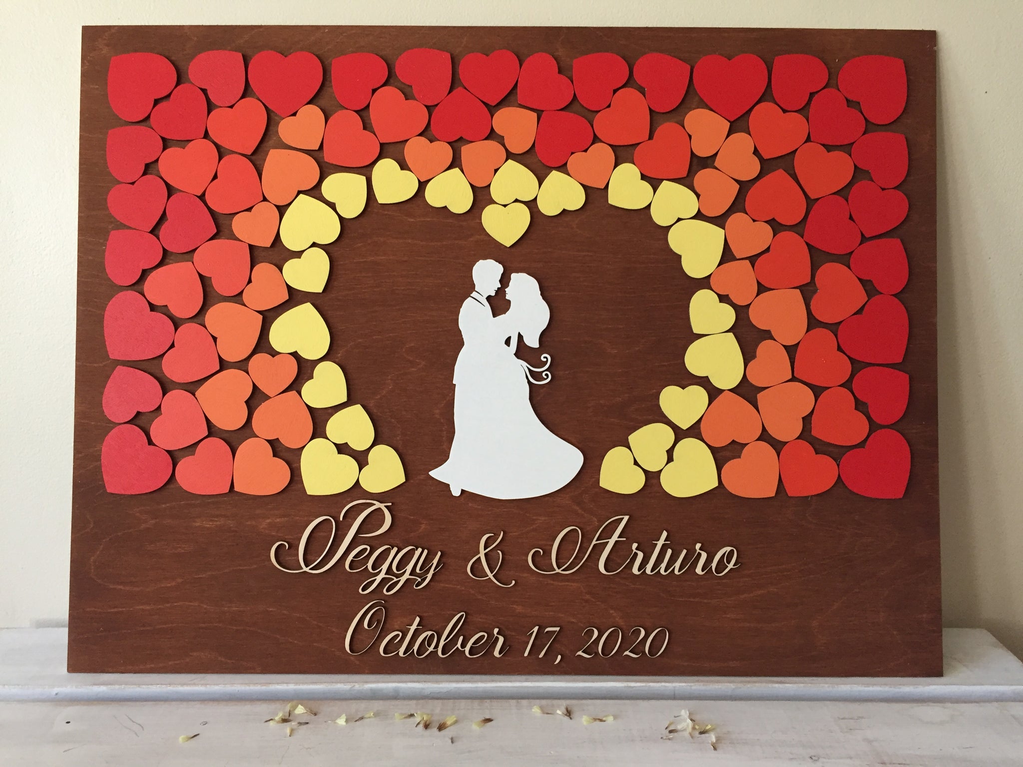 Guest book alternative with embracing couple in the centre and surrounded by hearts that also form a heart shape around the couple. The hearts are painted in fall colors from bright red to orange to yellow and there is a 3D cutout of the names of the couple in calligraphic font. The date is just below the names.