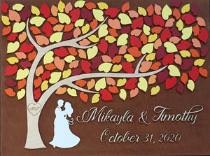 A wood guest book alternative made in a fall themed colors with personalized names, date and initials in a heart in the tree trunk and a couple silhouette underneath the tree.